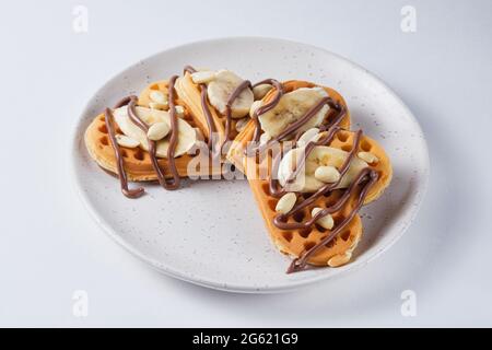 Waffle belgi dolci per colazione su un piatto bianco. Waffle con arachidi, banana e cioccolata. Foto Stock