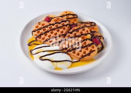 Waffle belgi dolci per colazione su un piatto bianco. Waffle con yogurt, lamponi, cioccolato e sciroppo di pesca. Piatto su un piatto bianco. Foto Stock