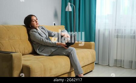 giovane donna in un abito formale si siede sul divano togliendo le scarpe. Massaggio ai piedi, gonfiore ai piedi. Foto Stock