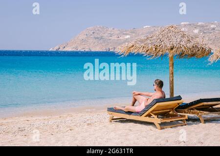 Elia Beach Mykonos, un giovane ragazzo sulla spiaggia di Mykonos durante l'estate con un ombrellone e sedie da spiaggia di lusso letti, l'oceano blu a Elia spiaggia Mykonos Grecia. Foto Stock