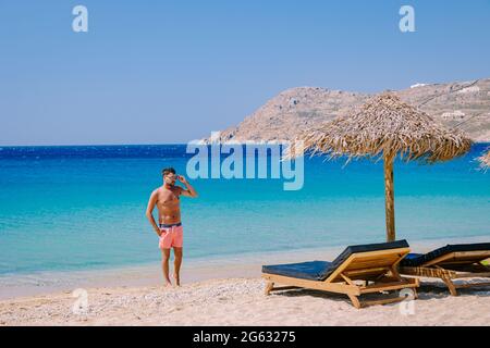 Elia Beach Mykonos, un giovane ragazzo sulla spiaggia di Mykonos durante l'estate con un ombrellone e sedie da spiaggia di lusso letti, l'oceano blu a Elia spiaggia Mykonos Grecia. Foto Stock