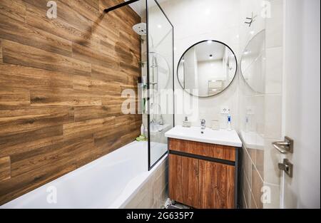 Versione elegante e confortevole del design del bagno in un piccolo spazio. Colore bianco della vasca rettangolare e del lavabo in armoniosa combinazione con specchio rotondo integrato da decorazioni in legno. Foto Stock