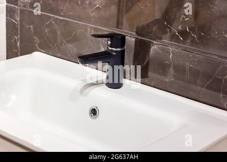 Nuovo rubinetto d'acqua nero moderno con lavello bianco sullo sfondo della parete in ceramica del bagno. Foto Stock