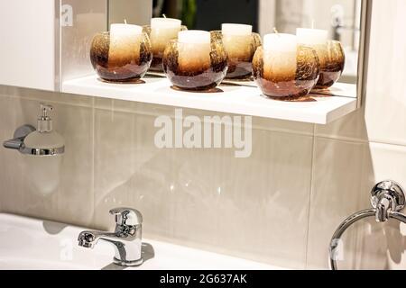 Nuovo rubinetto d'acqua in metallo moderno con lavello bianco e candele sullo sfondo della parete in ceramica del bagno. Foto Stock