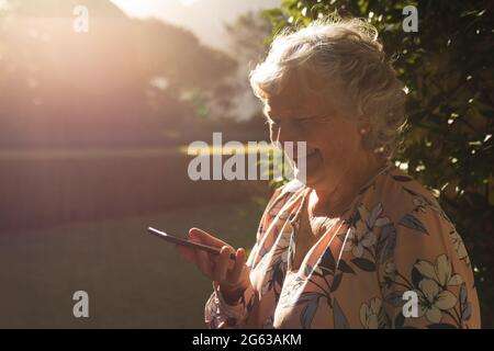 Sorridente donna caucasica senior utilizzando smartphone in giardino soleggiato Foto Stock