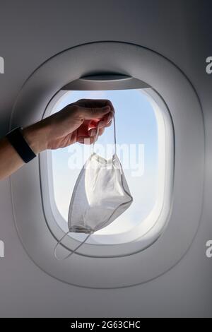 Mano che tiene la maschera protettiva contro il finestrino dell'aeroplano. Temi che viaggiano in una nuova protezione normale e personale durante la pandemia covid-19 Foto Stock