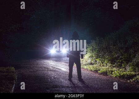 Una figura con cappuccio spoky, silhouette contro i fari di un'auto. Su una strada di campagna piovosa di notte. Foto Stock