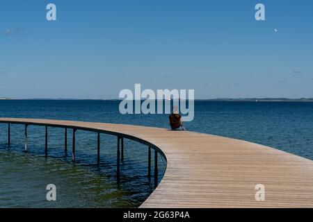 Una donna snella si siede da sola su una passerella circolare che conduce fuori e sopra un oceano blu calmo Foto Stock