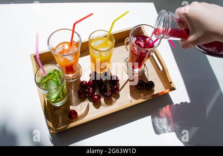 Un vassoio di legno con bevande colorate: Limonata arancione, gialla e cetriolo. Nelle vicinanze si trovano le ciliegie. Una mano versa il succo rosso da un decanter in un bicchiere. C Foto Stock