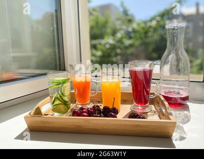 Vassoio di legno con bevande colorate, ciliegie su un davanzale. Decanter con i resti di succo rosso. Finestra aperta, estate calda, luce solare. Foto Stock