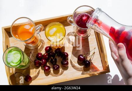 Un vassoio di legno con bevande colorate: Limonata arancione, gialla e cetriolo. Nelle vicinanze si trovano le ciliegie. Una mano versa il succo rosso da un decanter in un bicchiere. C Foto Stock