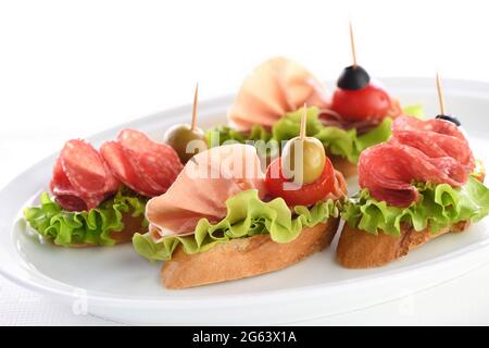 Tartine di baguette tenue con lattuga, salame o prosciutto di Parma, pomodori, mozzarella e olive. Piatto assortito di prelibatezze per la festa. Foto Stock