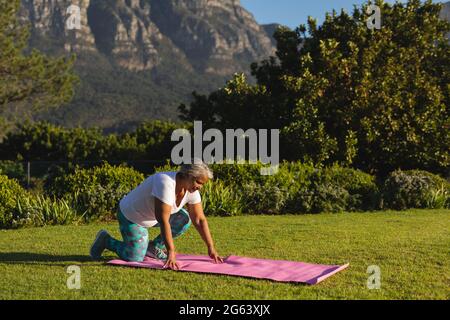 Donna afro-americana anziana inginocchiata sul tappeto yoga sull'erba in una splendida campagna Foto Stock