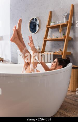 donna gioiosa con gambe rialzate che prendono il bagno in vasca con schiuma da bagno Foto Stock