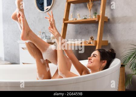 donna allegra con gambe rialzate che prendono il bagno nella vasca da bagno con schiuma da bagno Foto Stock