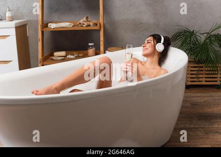 donna felice in cuffie ascoltare musica e tenere un bicchiere di champagne mentre si prende il bagno Foto Stock