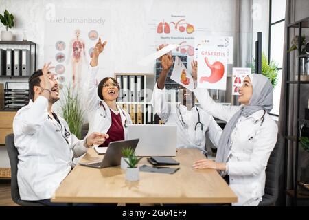 Gruppo di quattro medici specialisti diversi che gettano su carte dopo lunghe e difficili discussioni in ufficio. Riunitosi con successo. Cooperazione internazionale. Foto Stock