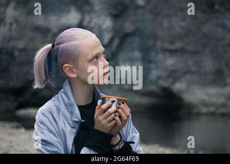 ritratto di una ragazza adolescente che riposa con una tazza sulla riva del fiume guardando le rocce costiere Foto Stock