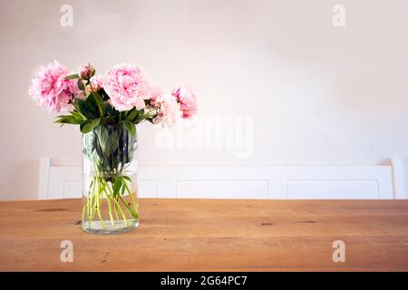 Un mazzo di color rosa pastello con foglie verdi in vaso di vetro sul tavolo di legno vicino alla tessitura bianca delle pareti, accogliente decorazione luminosa per una casa elegante Foto Stock