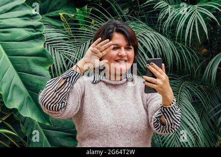 Donna che saluta la sua famiglia durante una videochiamata sul suo cellulare all'aperto su sfondo verde. Concetto di telecomunicazione Foto Stock