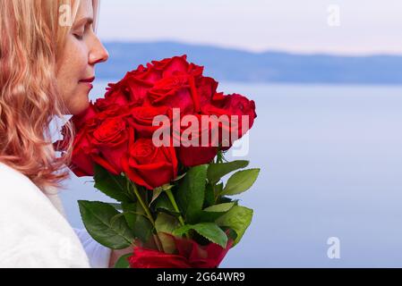 Donna felice in rosa bianca con un bouquet di rose rosse guarda dal balcone dell'hotel sul mare Foto Stock