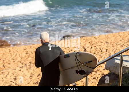 Il surfista di mezza età con muta porta la tavola da surf alla spiaggia, Sydney, NSW, Australia Foto Stock