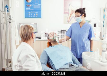 Assistente ortodontista che mette la maschera di ossigene al paziente che analizza il dolore dei denti durante l'appuntamento stomatologico. Team medico che lavora in ambulatorio di odontoiatria controllo dente preparazione per la chirurgia Foto Stock