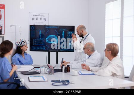 Neurologo che mostra la radiografia digitale ai colleghi medici che analizzano la presentazione delle malattie cerebrali utilizzando l'alta tecnologia nella sala riunioni. Il team ospedaliero sta analizzando il trattamento malato esaminando le competenze sanitarie Foto Stock