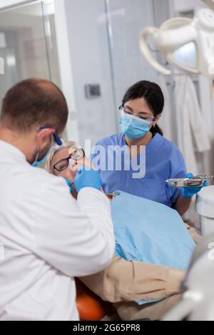 Dentista con assistente installare l'impianto nella clinica moderna della moutina del paziente. Donna anziana durante l'esame medico con stomatolog in studio dentistico con attrezzature arancioni. Foto Stock