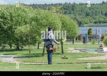 BIELORUSSIA, MINSK - 15 GIUGNO 2021: Un uomo con un tosaerba portatile taglia l'erba in un parco cittadino. Fotografia di scorta. Foto Stock