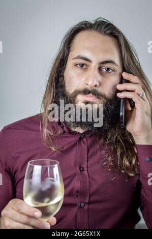 Ritratto di un giovane uomo d'affari mediorientale con una barba e capelli lunghi che tiene un bicchiere di vino bianco mentre si ha una conversazione telefonica Foto Stock