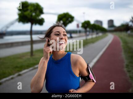 Donna attiva sorridente eserizzante all'aperto. Atleta giovane che usa l'auricolare e che corre il eriver besid di jogging Foto Stock