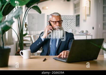 Uomo d'affari senior che chiama con il telefono cellulare mentre lavora con il computer portatile all'interno di un ufficio moderno - Focus on Face Foto Stock