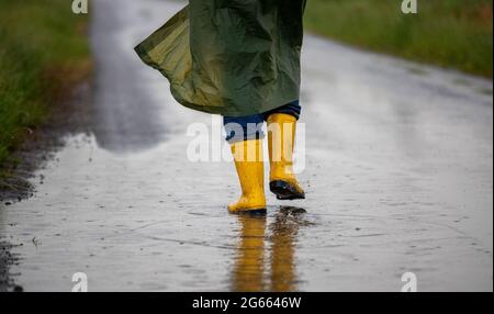 Primo piano delle gambe del contadino in stivali gialli e impermeabile verde che cammina sulle puddddle dopo la pioggia in campo Foto Stock