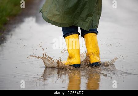 Primo piano delle gambe del contadino in stivali gialli e impermeabile verde che salta sulle puddddle dopo la pioggia in campo Foto Stock