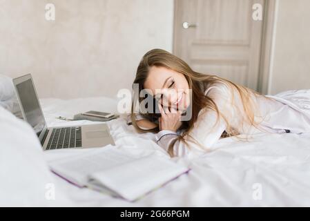 Foto di giovane donna allegra in pigiama digitando su laptop e sorridenti mentre è seduto sul letto in camera di luce Foto Stock