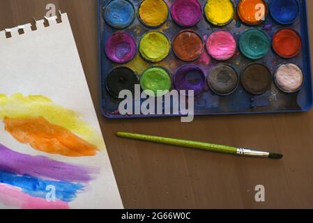 Un pezzo di carta bianca con colori dipinti di un bambino accanto a una scatola di vernice con vernici ad acquerello e un pennello Foto Stock