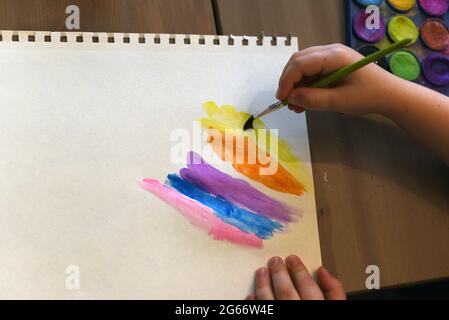 Un bambino usa un pennello per dipingere un pezzo di carta bianca con colori dipinti accanto a una scatola di vernice con vernici ad acquerello Foto Stock