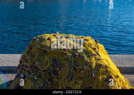 Le reti da pesca si accatastano sul molo del porto di fisher che si asciugano sotto il sole. Reti da pesca di colore giallo con corde e galleggianti su fondo di mare increspato. Isola greca C Foto Stock