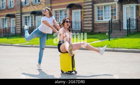 Tre allegri amici caucasici si divertono e si inseguono mentre cavalcano una valigia. Le donne eccitate vanno in viaggio. La bionda brunette e. Foto Stock