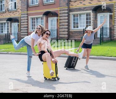 Tre allegri amici caucasici si divertono e si inseguono mentre cavalcano una valigia. Le donne eccitate vanno in viaggio. La bionda brunette e. Foto Stock