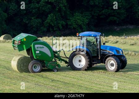 L'agricoltore che fa grandi balle rotonde di grandine con un trattore New Holland TS135 e una pressa McHale F5500, estata sera a Wensleydale, North Yorkshire Foto Stock