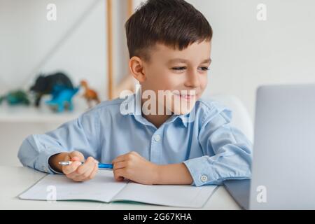 Ragazzino intelligente che fa i compiti usando il laptop e Internet. E-learning scuola elementare tramite videochiamata. Ragazzo che studia online a casa Foto Stock