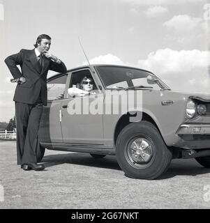 1975, storico, su un'area pavimentata fuori strada, un uomo in tuta in piedi da un'auto a motore Datsun Cherry 100A, Inghilterra, Regno Unito, con una donna in occhiali da sole seduto al posto di guida. Si noti il grande reverel della giacca da uomo e il fondo svasato dei pantaloni e le scarpe con plateau, gli stili di moda distintivi degli anni '70s. La società automobilistica giapponese Nissan ha iniziato ad esportare nel Regno Unito automobili con marchio Datsun nel 1968 e il marchio ha avuto un grande successo, con auto affidabili e a buon prezzo, in un momento in cui l'industria automobilistica britannica era in crisi, con scioperi della forza lavoro e automobili di uno stile, qualità e affidabilità scadenti. Foto Stock