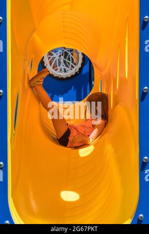 Giovane ragazza in vista laterale rilassarsi all'interno di un tubo di plastica gialla o tubo sul parco giochi arrampicata telaio e scivolo Foto Stock