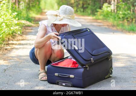 Una giovane bella ragazza apre una valigia con le cose su una strada forestale sullo sfondo di verde fogliame in una giornata estiva di sole. Messa a fuoco selettiva Foto Stock