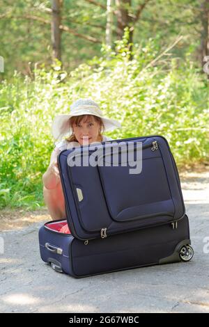 Una giovane bella ragazza apre una valigia con le cose su una strada forestale sullo sfondo di verde fogliame in una giornata estiva di sole. Messa a fuoco selettiva Foto Stock