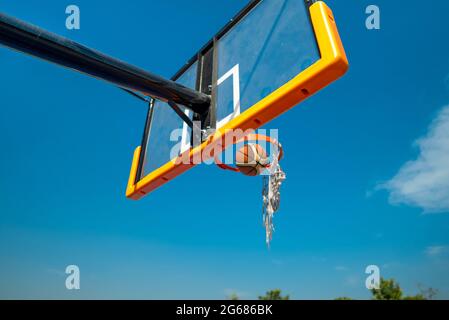 Palla di pallacanestro che cade attraverso rete rotta sul parco giochi dopo il colpo. Cielo blu su sfondo Foto Stock