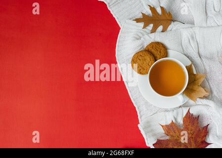 Il concetto di una colazione autunnale: Una tazza di tè, biscotti d'avena, foglie gialle cadute su una coperta bianca lavorata su uno sfondo rosso. Spazio di copia, fl Foto Stock