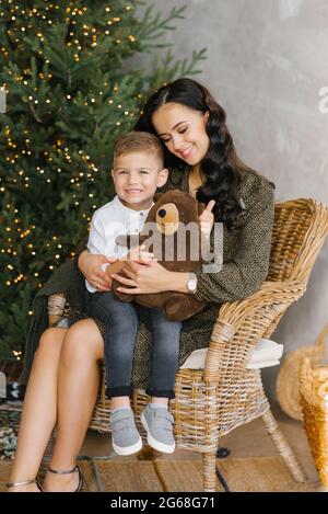 Ritratto di una madre felice con il suo figlio di quattro anni, che è seduto in grembo su una sedia in vimini sullo sfondo di un albero di Natale Foto Stock